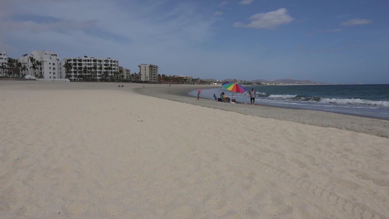 Wide San Jose del Cabo beach view with pale sand in front, a rainbow umbrella and small figures near the surf, white buildings at left, and blue ocean at right.