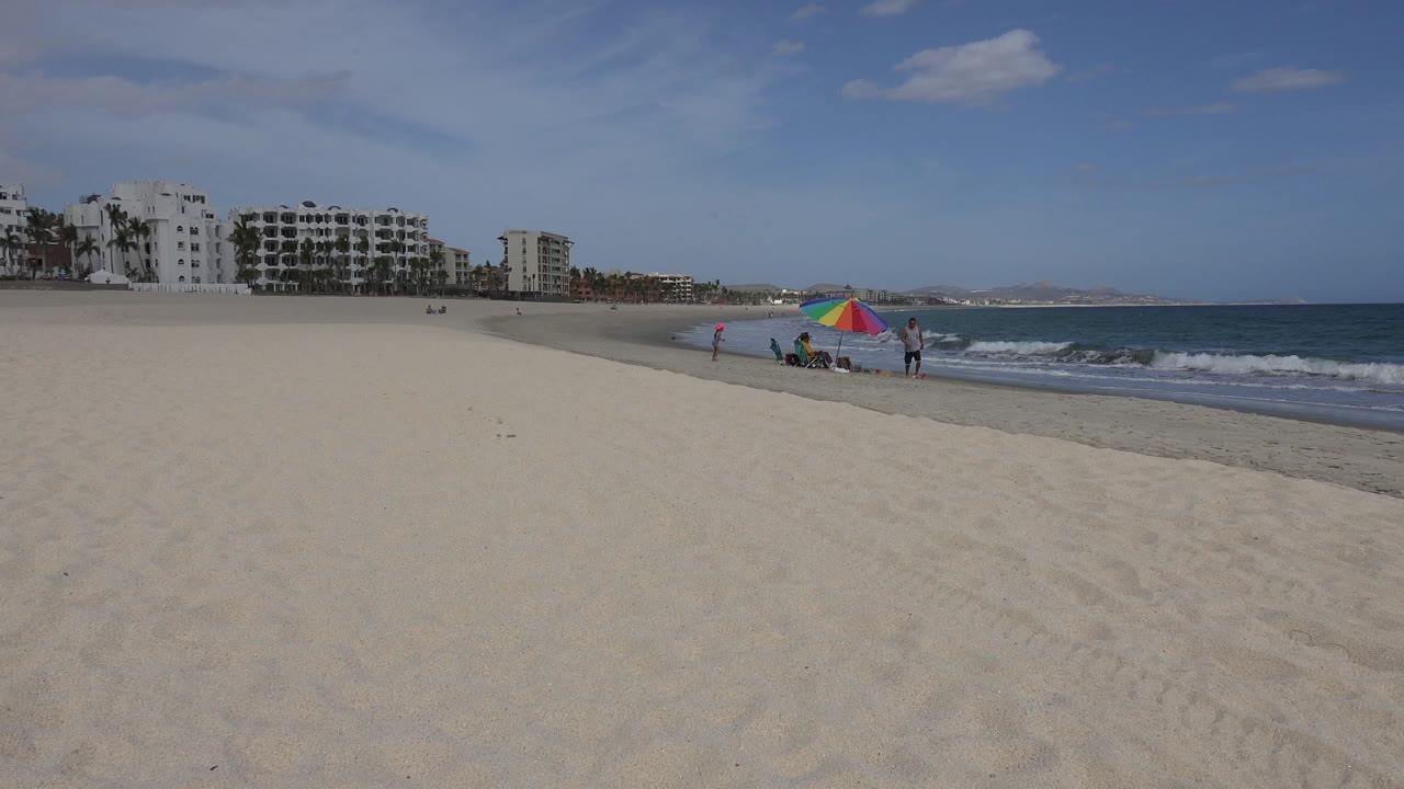 San Jose del Cabo shoreline with a large empty sand foreground, small people beside a multicolor umbrella, low waves on the right, and waterfront buildings in the distance.
