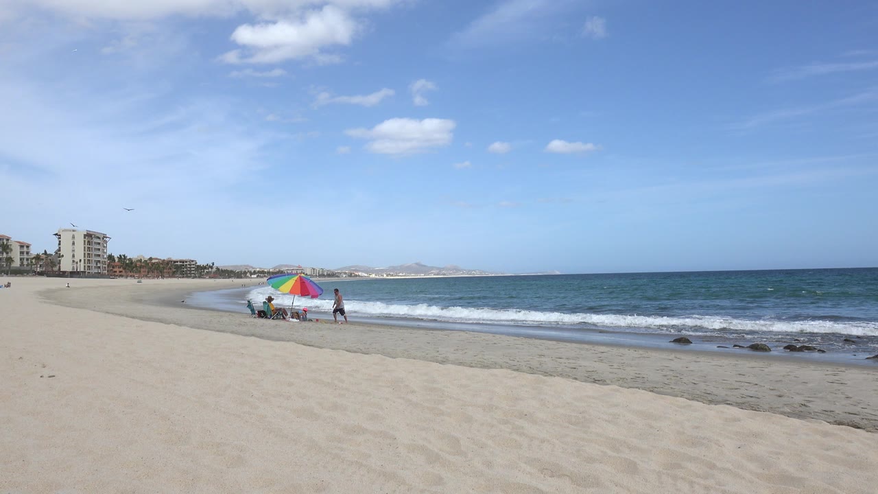 San Jose del Cabo beach view with pale sand in the foreground, blue ocean at right, distant buildings at left, and small beachgoers near a multicolor umbrella.