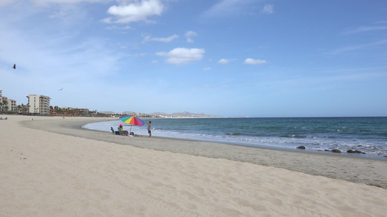 San Jose del Cabo beach view one second in, with the umbrella slightly right of the distant buildings, two small people by the waterline, and a low wave band crossing the shore.