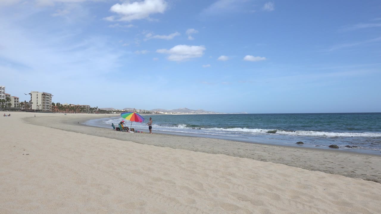 Wide San Jose del Cabo beach view with pale sand in front, blue ocean at right, a rainbow umbrella near the waterline, and a few distant people beside low buildings at left.