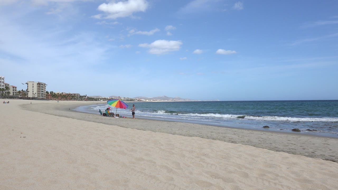 San Jose del Cabo shoreline with the rainbow umbrella slightly left of center, beach chairs on the sand, small waves rolling in, and distant people near the water.