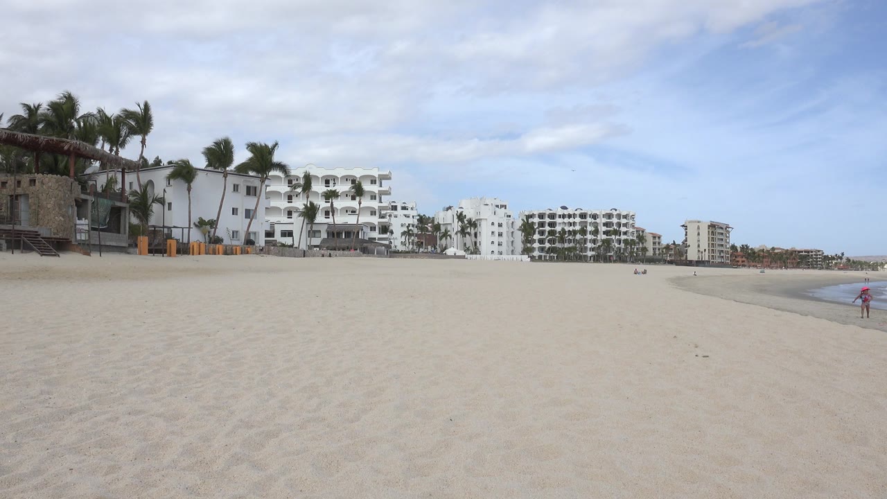 Wide sandy beach with white buildings, palms, and sea at right.