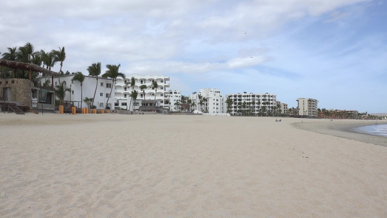 A wide San Jose del Cabo beach view shows open sand in the foreground, white beachfront buildings and palms in the background, sea along the right edge, and no visible people.