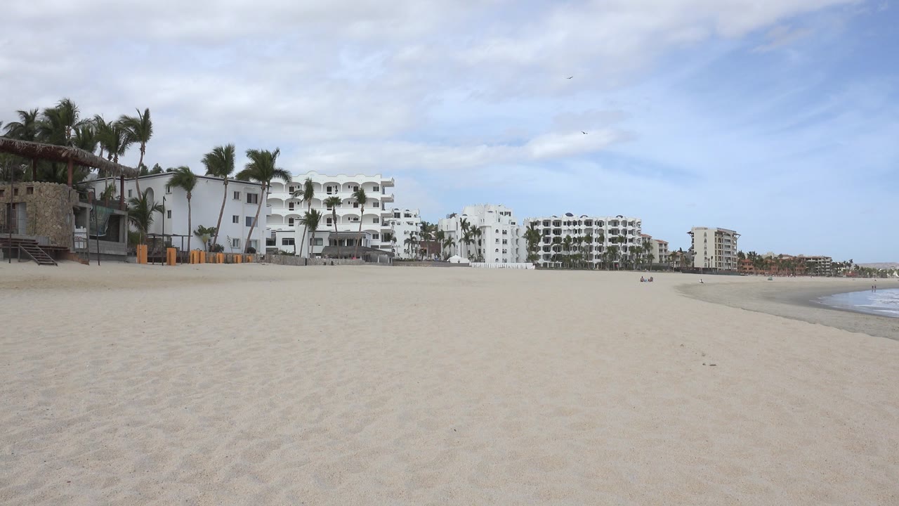The second San Jose del Cabo frame keeps the beach foreground broad, with white coastal buildings centered left, palms along the shore, a cloudy blue sky, and no visible people.