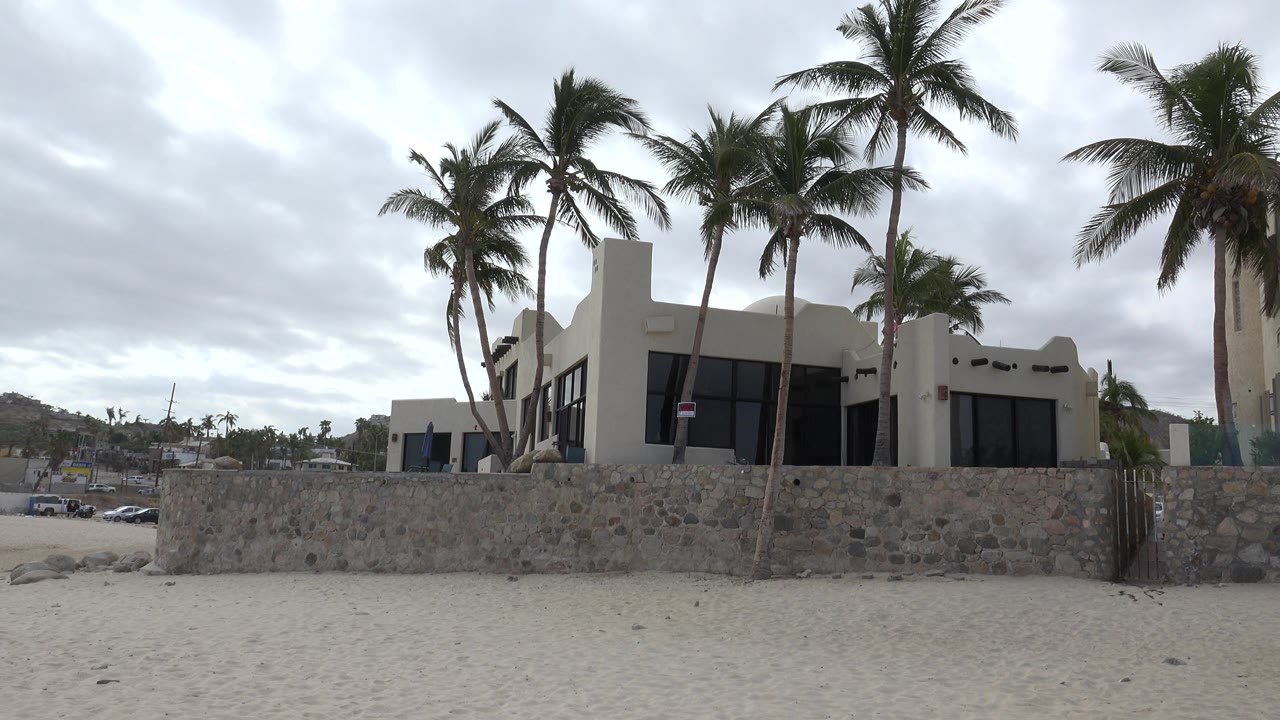 Beachfront house behind a stone wall with palm trees and sand