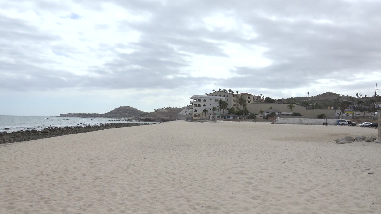 Wide sandy beach with ocean, rocks, hills, and coastal buildings under cloudy sky.