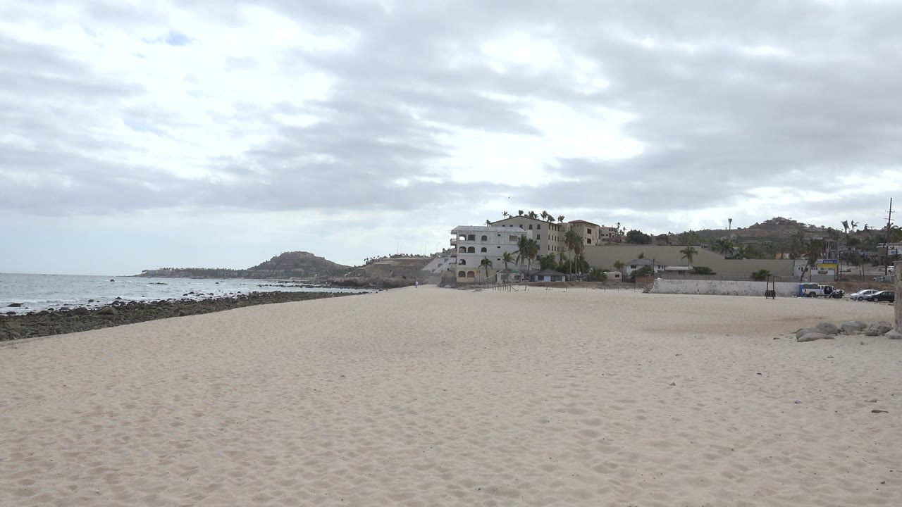 Pale sand stretches across the foreground in San Jose del Cabo, Mexico, with the ocean at left, a rocky edge near the water, white buildings beyond the beach, and no visible people.