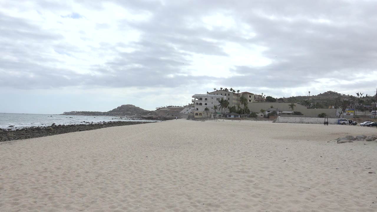 Wide sandy beach with rocky shoreline, ocean, buildings, and cloudy sky.