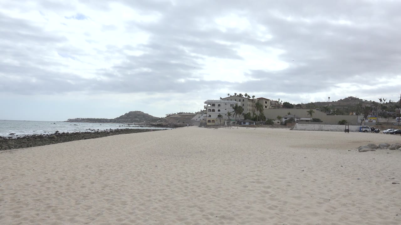 San Jose del Cabo shoreline view at one second, with the sandy beach dominating the foreground, small waves beyond the rocks, and white coastal buildings under a layered gray sky.