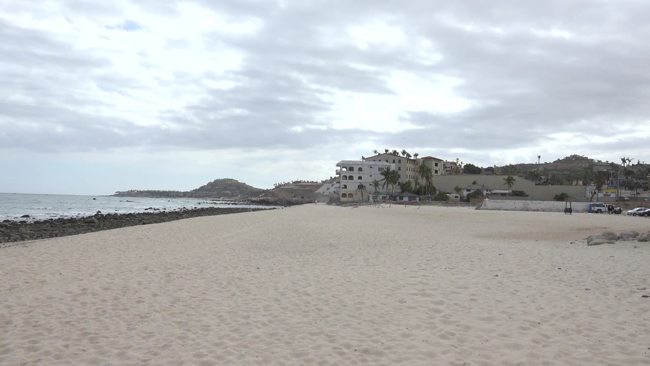 Empty sandy beach with ocean, rocks, distant building, hills, and cloudy sky.