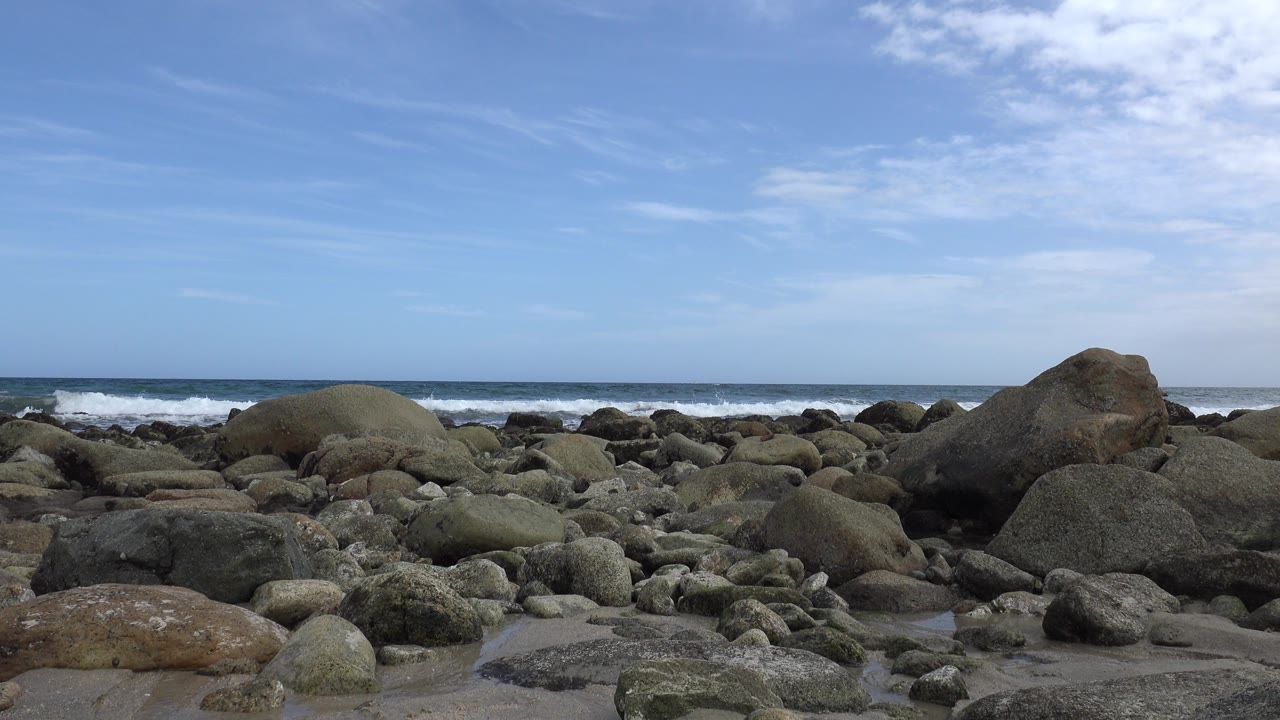 Rocky shoreline in San Jose del Cabo, Mexico with rounded gray stones, white surf on the horizon, blue sky above, and no visible people.