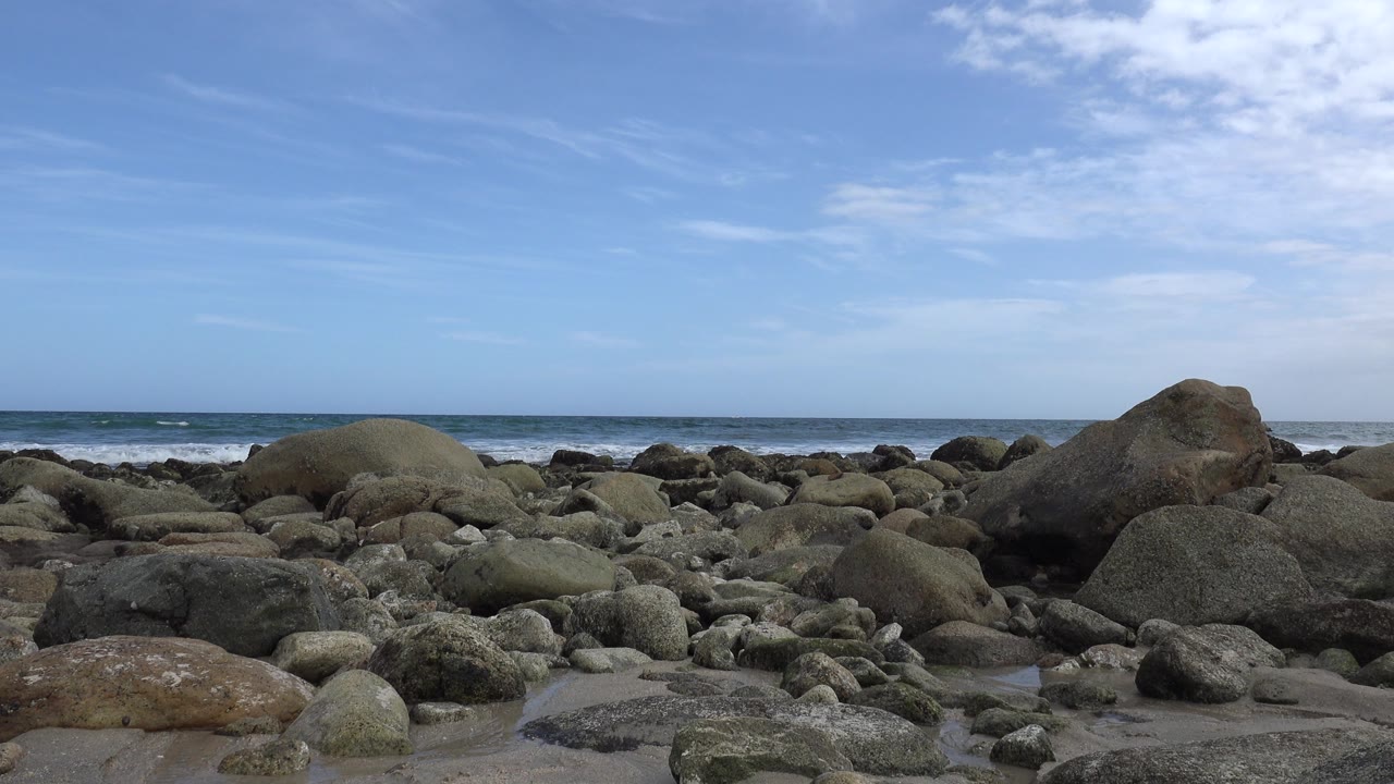San Jose del Cabo rocky beach with rounded stones in the foreground, low ocean waves on the horizon, blue sky with thin cloud, and no visible people.