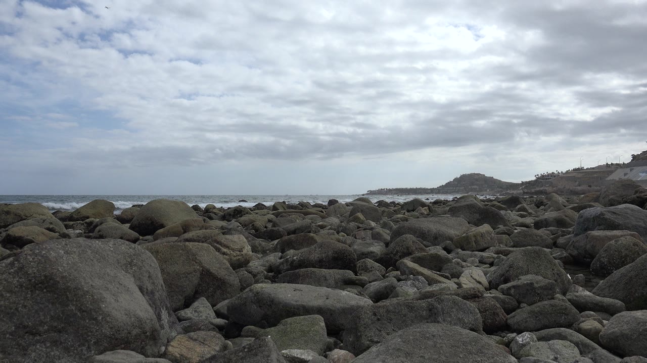 Opening frame of a rocky San Jose del Cabo shoreline with large foreground boulders, ocean surf on the left, distant headland on the right, and no visible people.