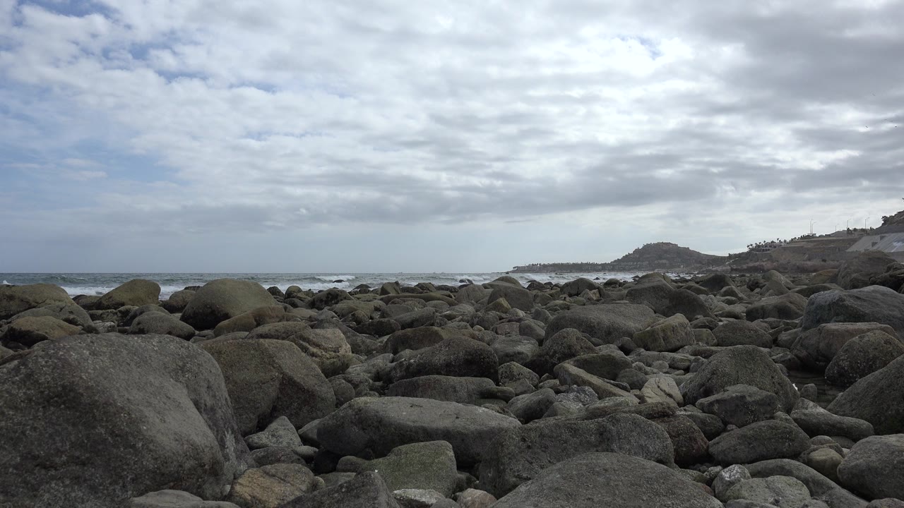 Opening frame of a rocky San Jose del Cabo shoreline with large boulders in front, surf beyond them, distant hills on the right, and no visible people.