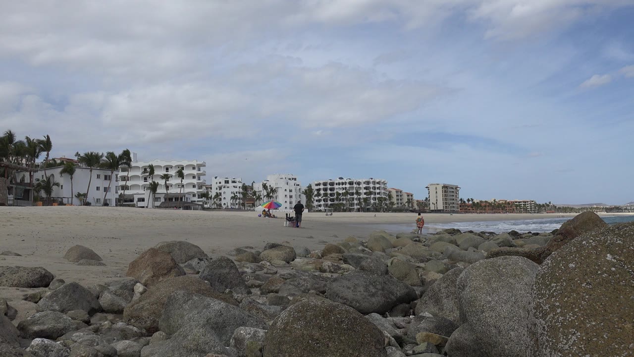Low beach view in San Jose del Cabo with rounded rocks in the foreground, pale sand, distant people, white hotel buildings, palms, and clouds over the shoreline.