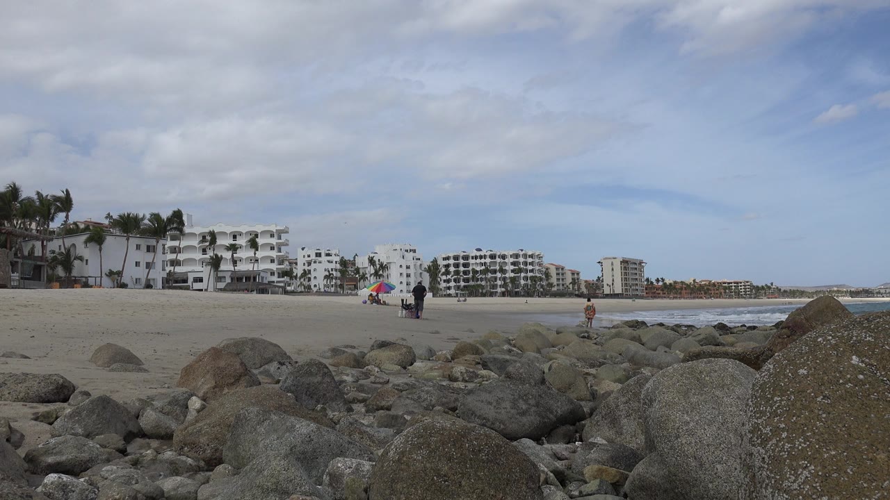 San Jose del Cabo beach frame with foreground boulders, a colorful umbrella near the sand, several small people, hotel blocks, palm trees, and waves along the right edge.