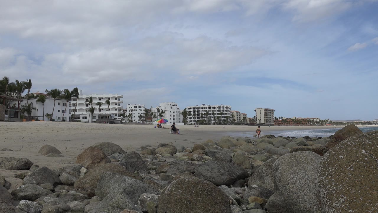Rocky foreground beside a sandy beach with distant people and beachfront buildings.