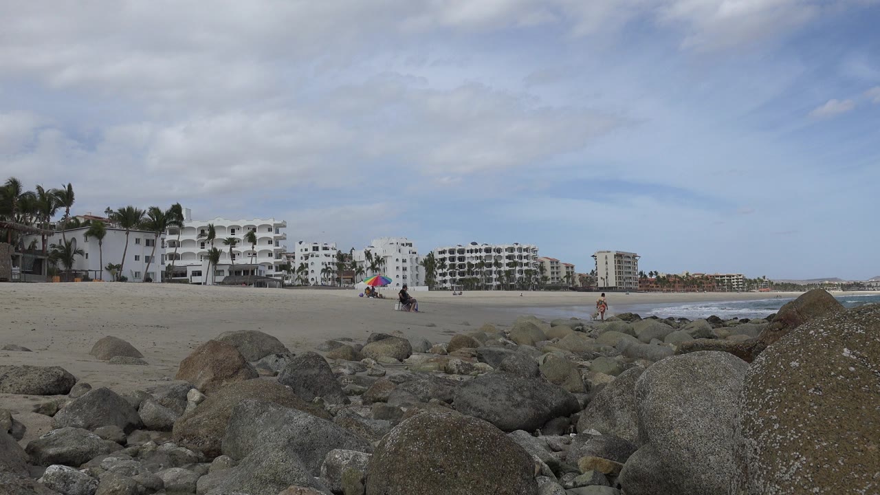 San Jose del Cabo beach view one second in, with foreground boulders, small people near a colorful umbrella, surf at right, and white buildings across the sand.