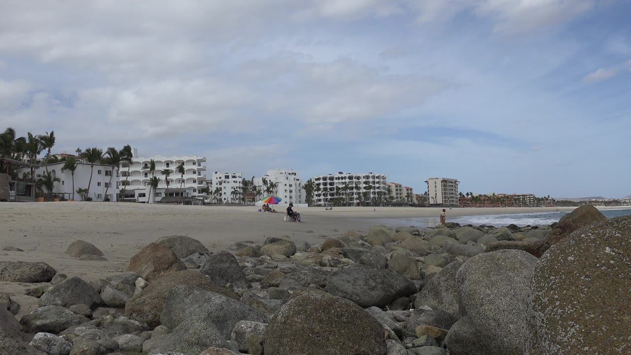 Opening frame of a San Jose del Cabo beach view with large foreground rocks, sand, distant waves, white buildings, palms, and tiny distant beachgoers.