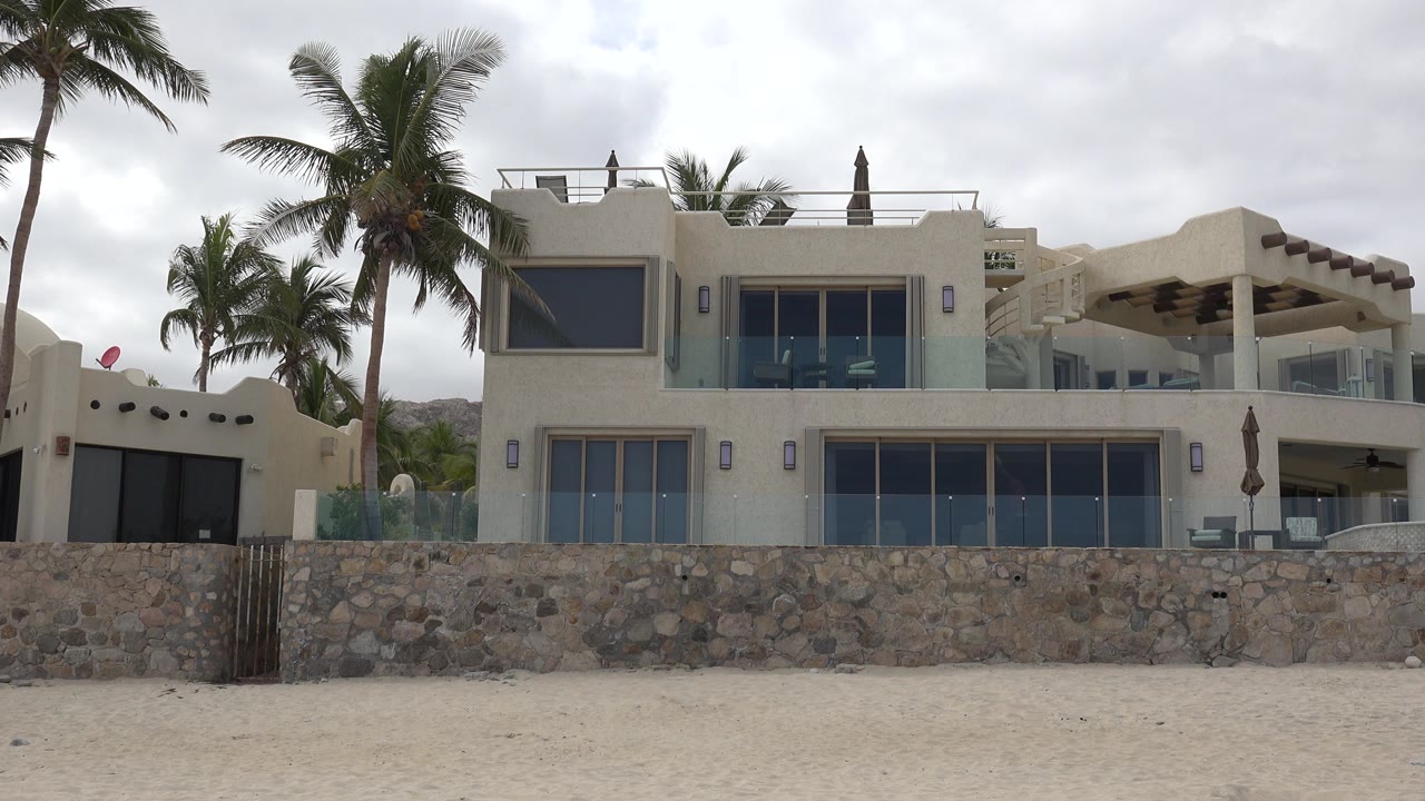 Opening frame of a white beachfront house in San Jose del Cabo, Mexico, behind a low stone wall with palms, sandy foreground, cloudy sky, and no visible people.