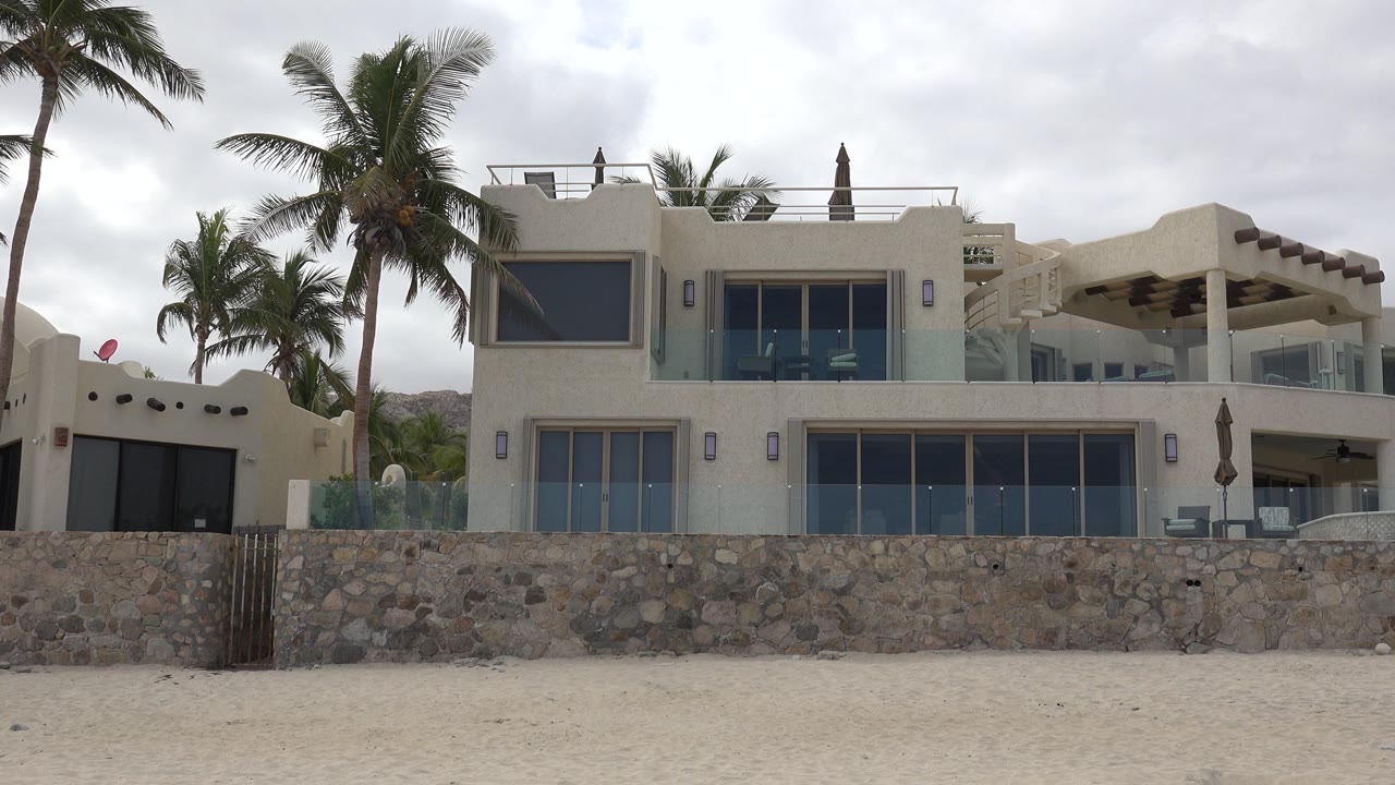 A steady view of the San Jose del Cabo beachfront house shows large glass doors, balcony railings, palm trunks, sand, and no visible people.