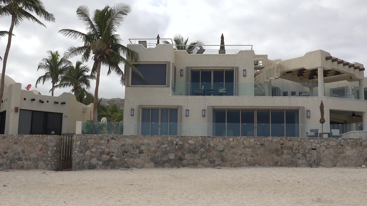 At 0:01, the San Jose del Cabo residential exterior remains centered, with the stone wall spanning the frame, palms rising on the left, and no visible people.