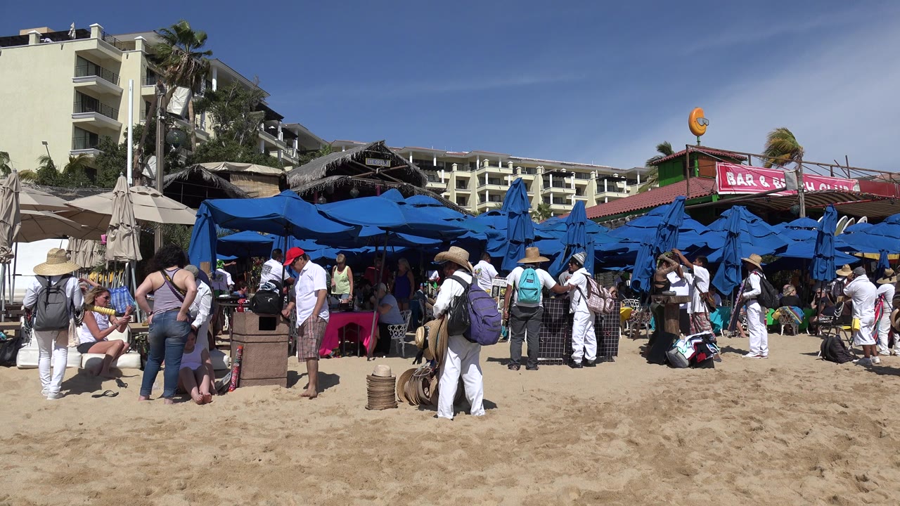 Busy sandy beach market with blue umbrellas and people gathered near buildings.