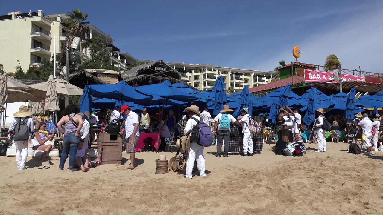 Cabo San Lucas beach crowd remains clustered around blue umbrellas, with a man in a red cap near the center, a person in a wide hat in the foreground, and buildings behind the market area.