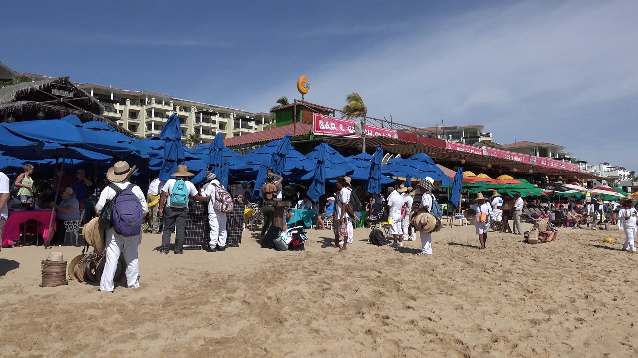 Cabo San Lucas beach scene with workers in hats gathered near bags and blue umbrellas while tourists stand along the sand under bright daylight.