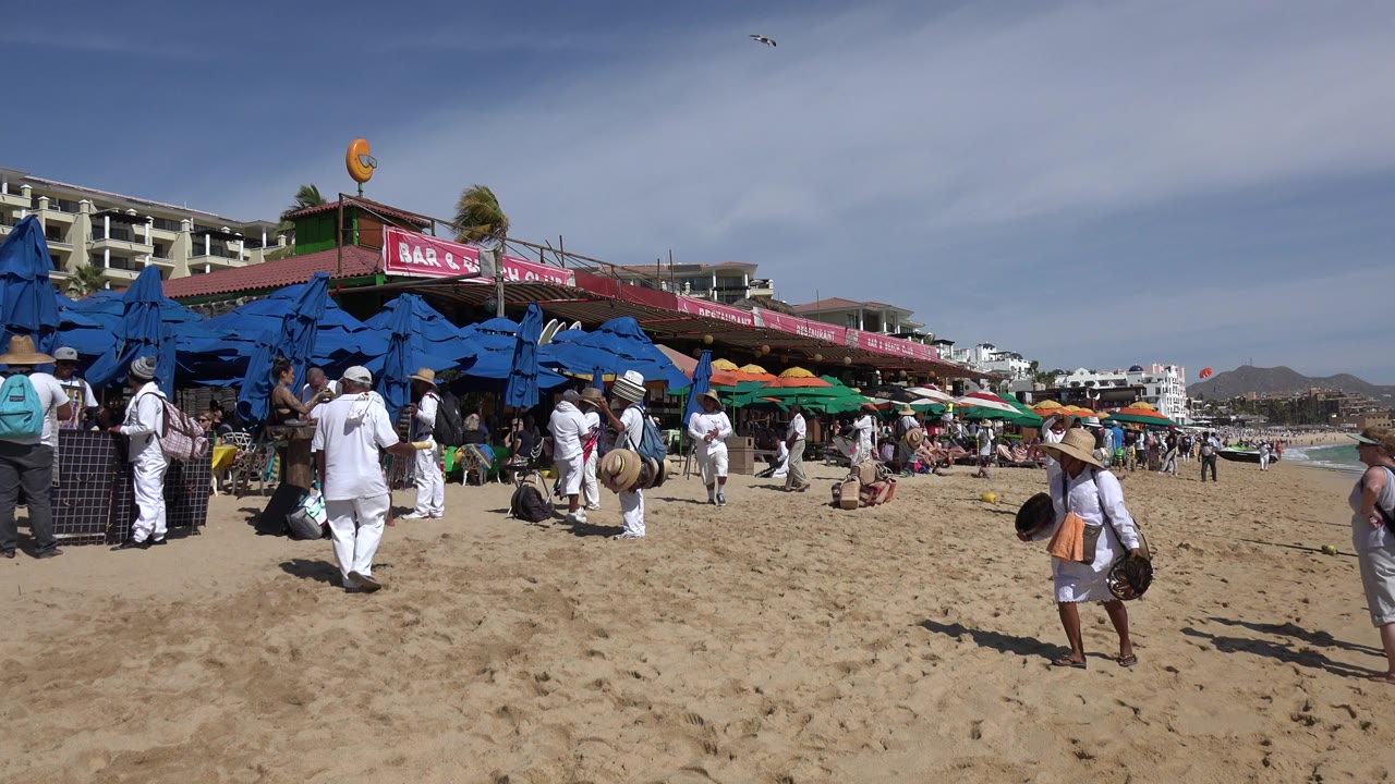 Crowded sandy beach with blue umbrellas and people walking near a beachfront bar sign.