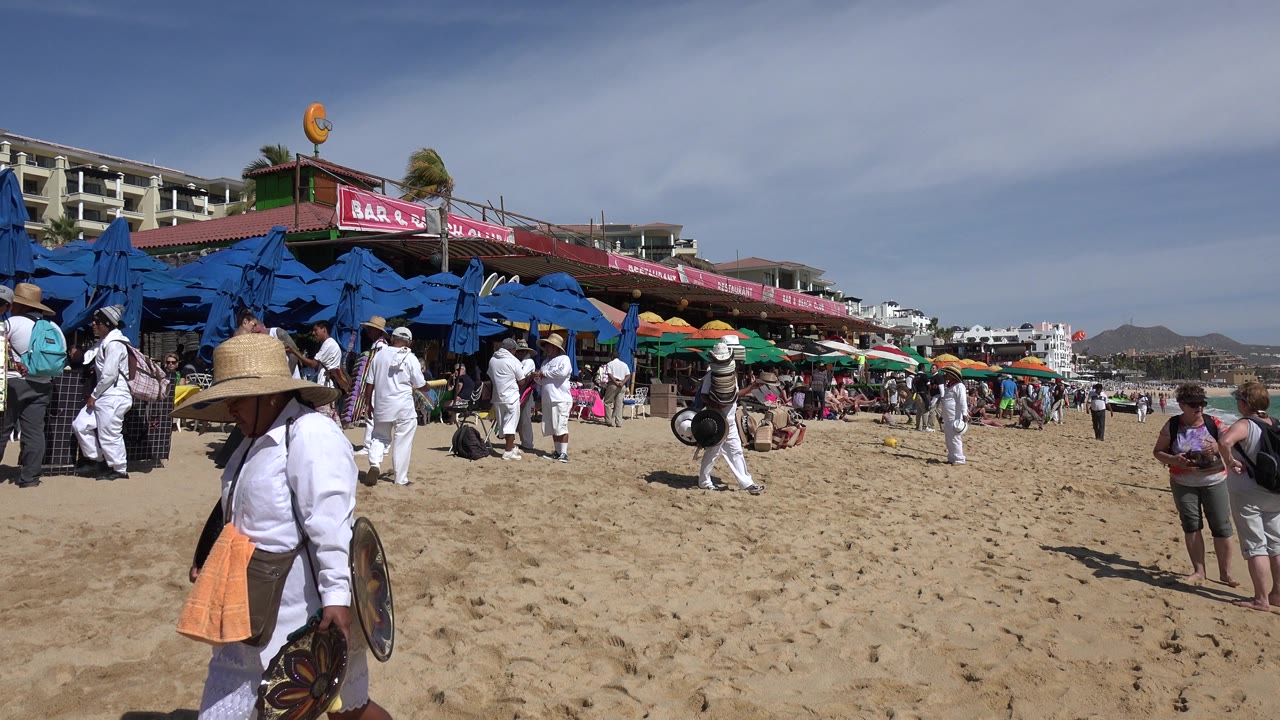 Beach crowd near blue umbrellas and a hat vendor on sand.