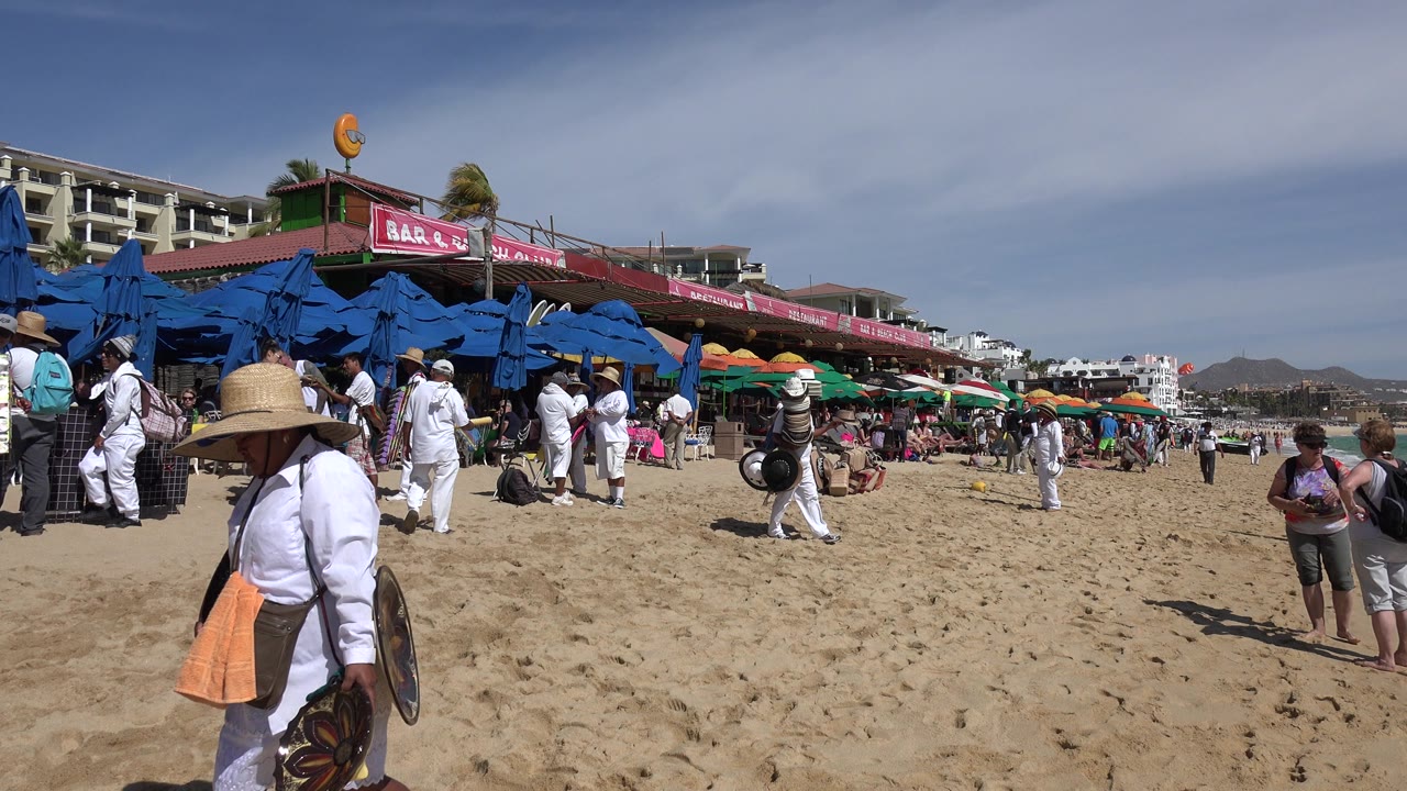 One second in, the Cabo San Lucas beach remains busy with the hat seller near the foreground and groups of people gathered under blue umbrellas by the shopfronts.