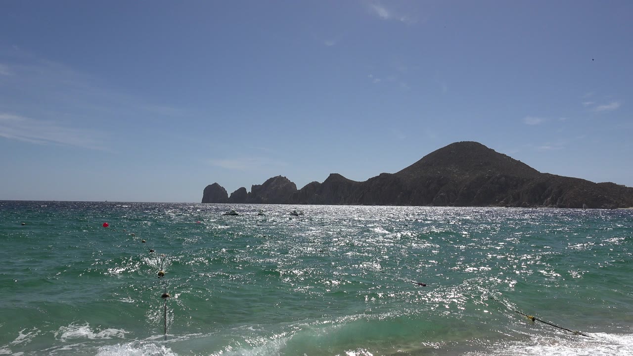 Cabo San Lucas ocean view with sunlit blue-green water, buoy markers near the shore, a dark rocky mountain range on the horizon, and no visible people.