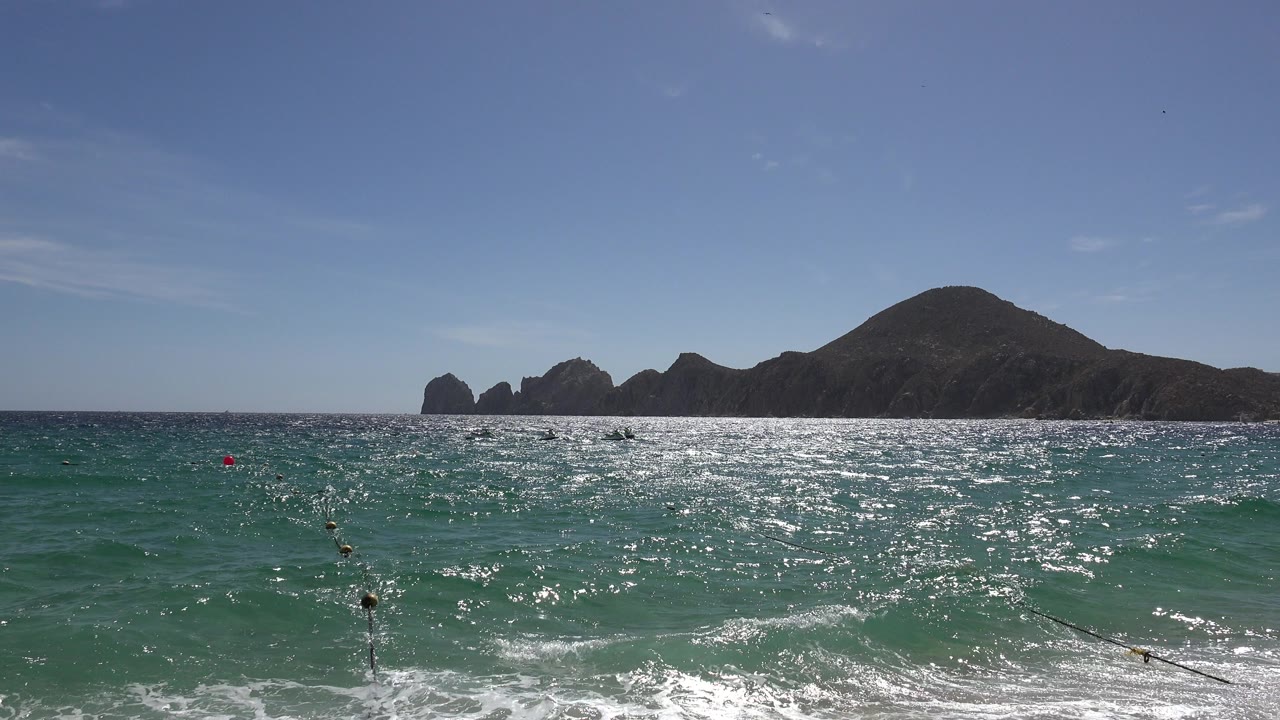 Cabo San Lucas beach-level frame with sparkling water across the midground, low surf at the bottom edge, buoy markers near the left side, and distant mountains under blue sky.