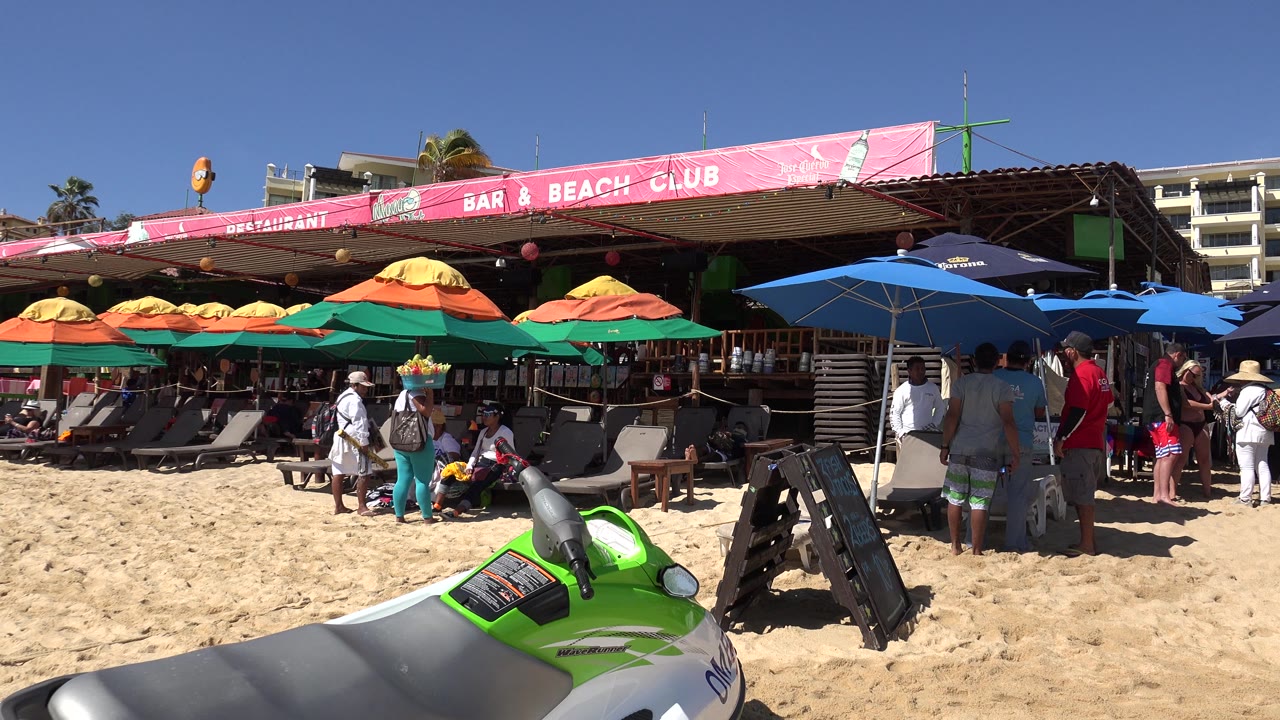 Sunny beach club frontage with tourists, umbrellas, signs, and watercraft.