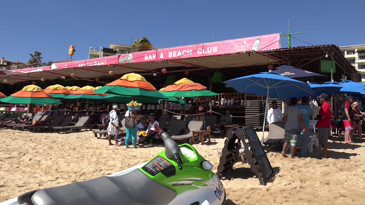 Cabo San Lucas beach club frame with the pink awning reading bar and beach club, shaded lounge chairs, several people near the entrance, and the watercraft cropped at the bottom.