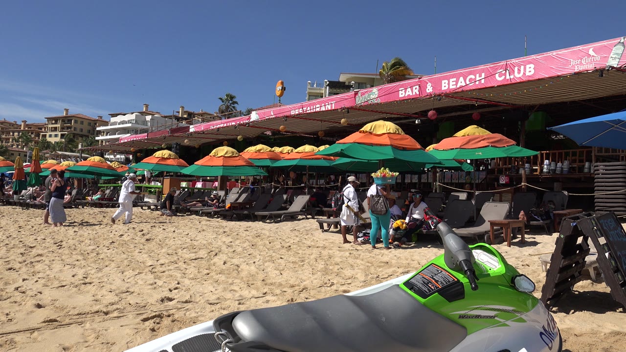 Busy sandy beach beside a bar and beach club with umbrellas and people.