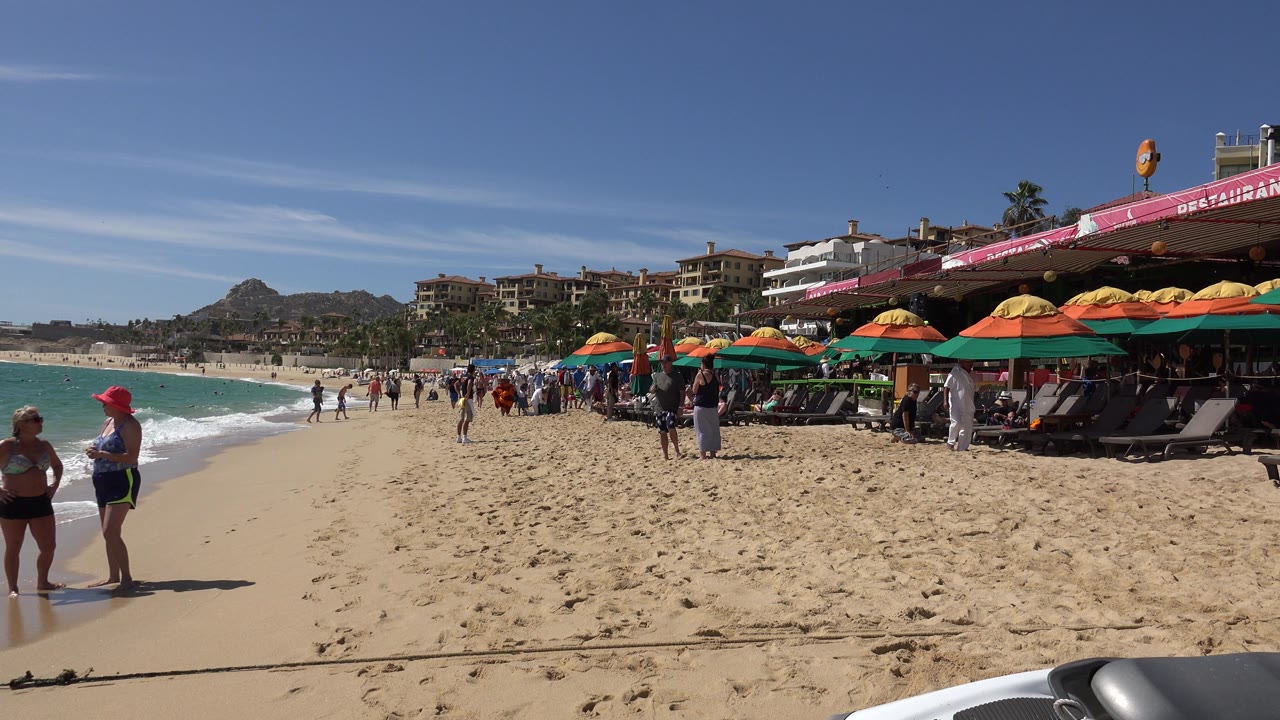 Two beachgoers stand near the surf in Cabo San Lucas while other tourists, umbrellas, lounge chairs, and hillside buildings fill the background.