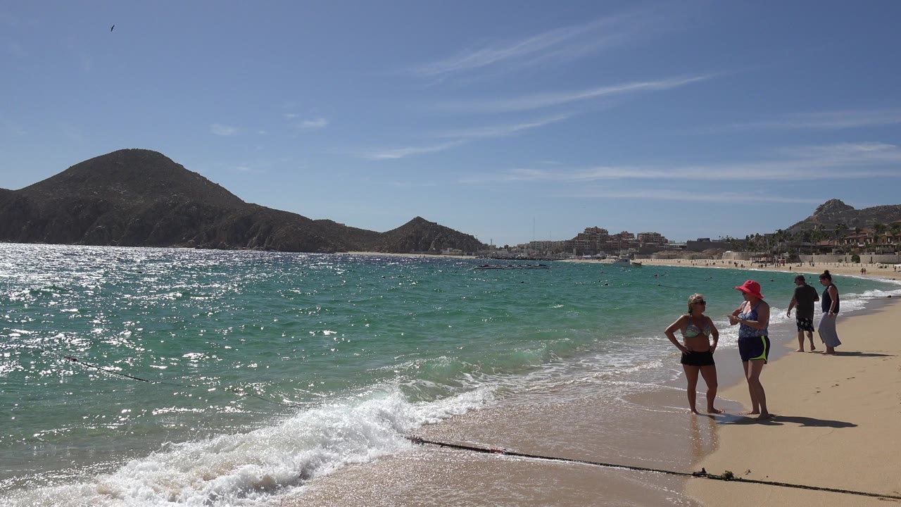 Cabo San Lucas beach scene with turquoise surf in the foreground, dark mountains across the bay, and several beachgoers standing near the waterline.
