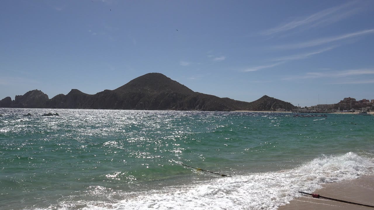 Cabo San Lucas surf rolls toward the foreground while strong sun reflection spreads across the water and dark hillside shapes line the background.