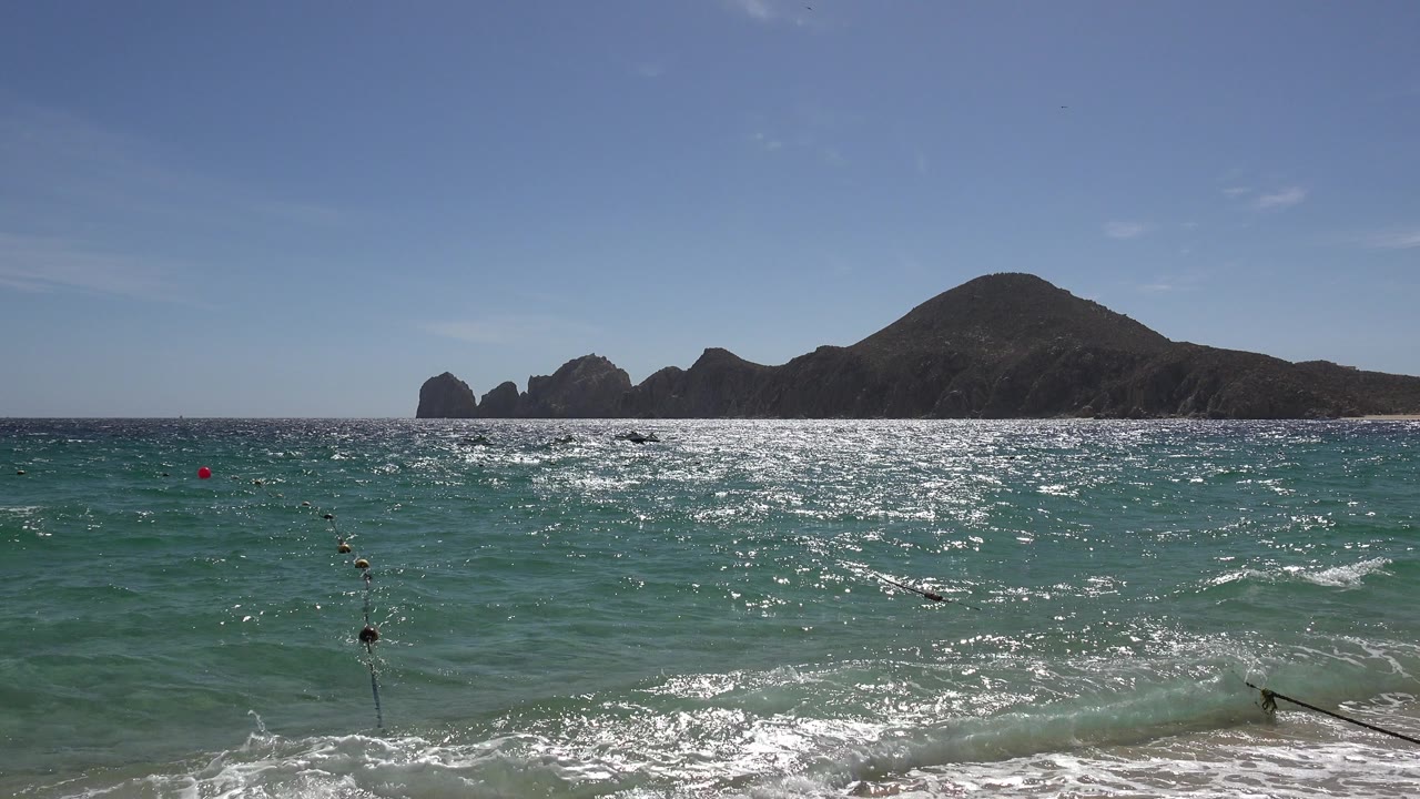 Cabo San Lucas ocean view with a rope or net line entering the lower foreground, sun glare on turquoise water, distant dark boulder formations, and no visible people.