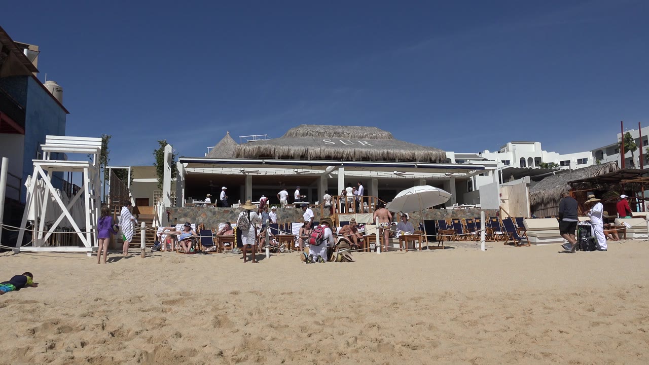 Wide sandy beach with people, umbrellas, chairs, and a beach building under blue sky.