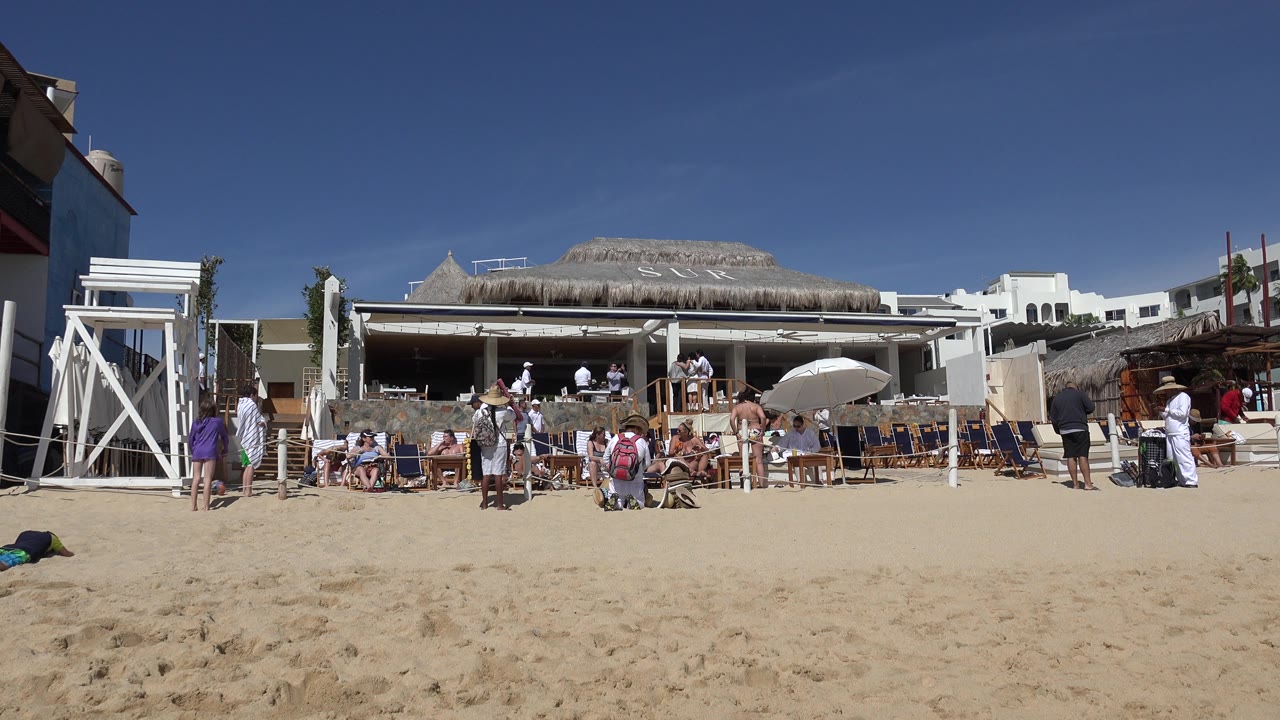 Wide ground-level view in Cabo San Lucas, Mexico showing people on a sandy beach, lounge chairs, umbrellas, a vendor in a hat and backpack, and white buildings behind them.