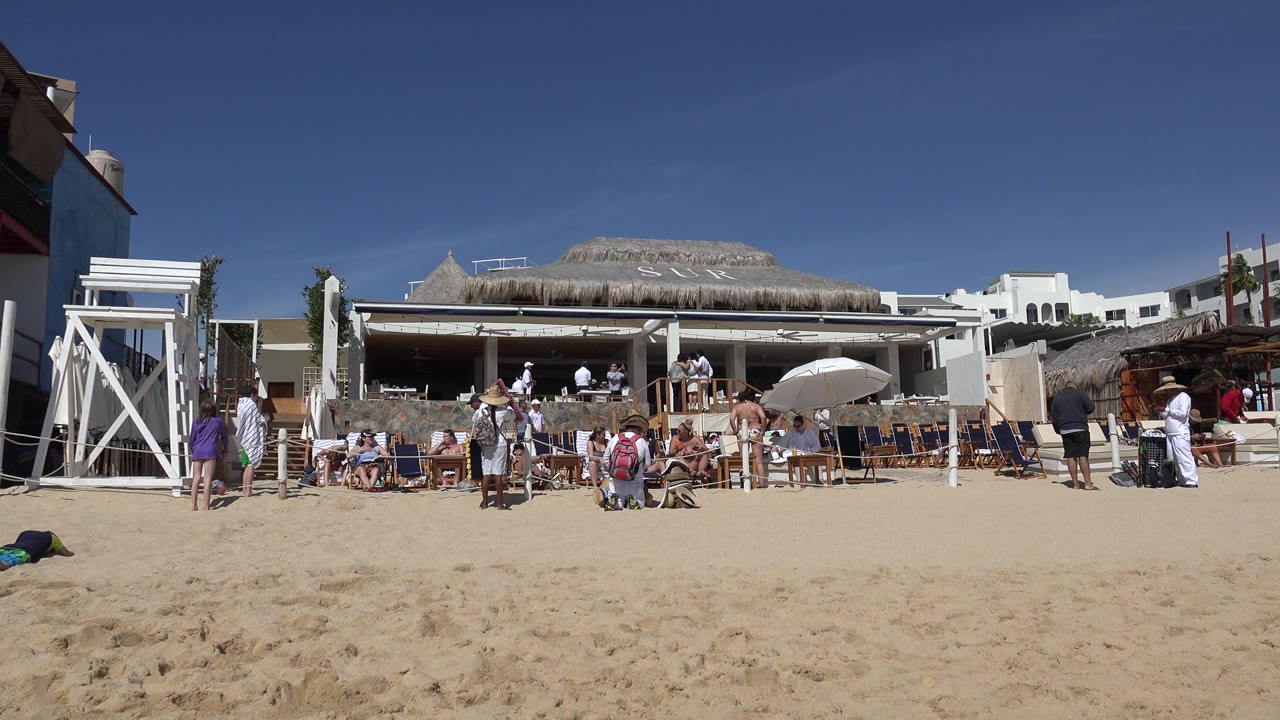 Cabo San Lucas beach crowd with seated tourists, standing people in white clothing, a hat-wearing vendor near the center area, and a thatched-roof structure in the background.