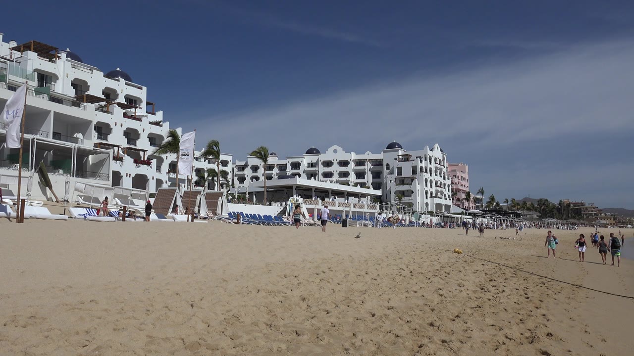Cabo San Lucas beach view with broad sand in front, white resort buildings and blue lounge chairs in the background, and a few distant people near the shoreline area.
