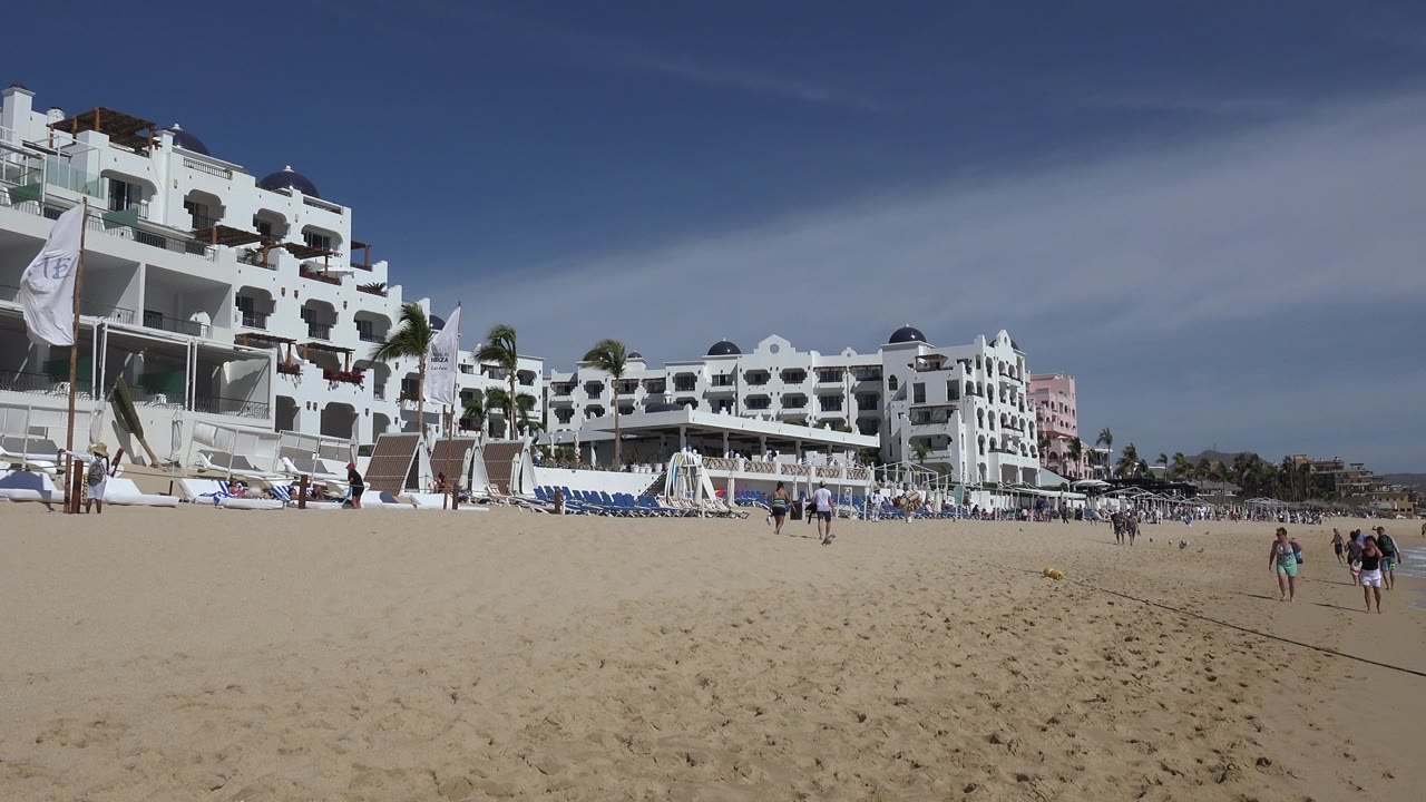Wide view of a Cabo San Lucas beach with open sand in front, white resort buildings to the left and center, lounge chairs, flags, and distant people under a blue sky.