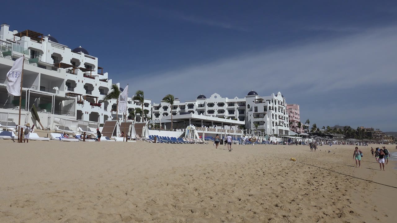 Cabo San Lucas beach scene with a broad sandy foreground, white hotel-style buildings, rows of blue beach chairs, and several people walking across the midground.