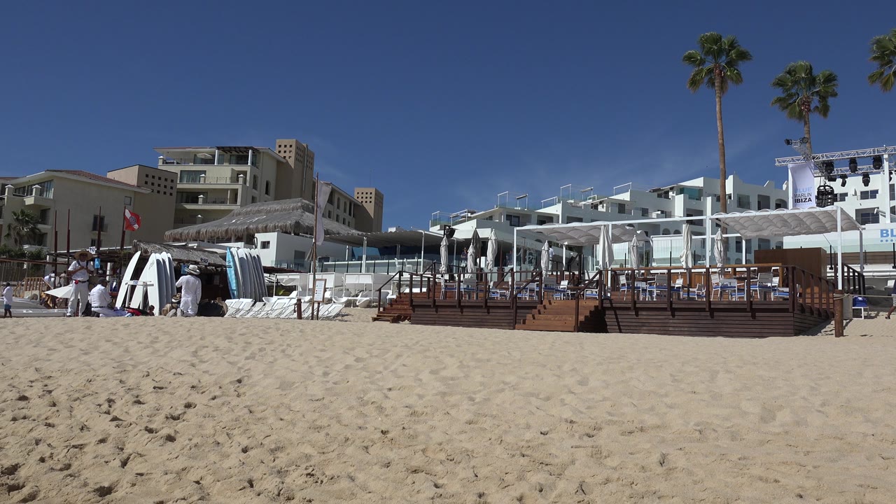 Cabo San Lucas beach resort scene with a raised wooden deck centered, rows of white lounge chairs to the right, resort buildings behind, and small distant people near the seating area.