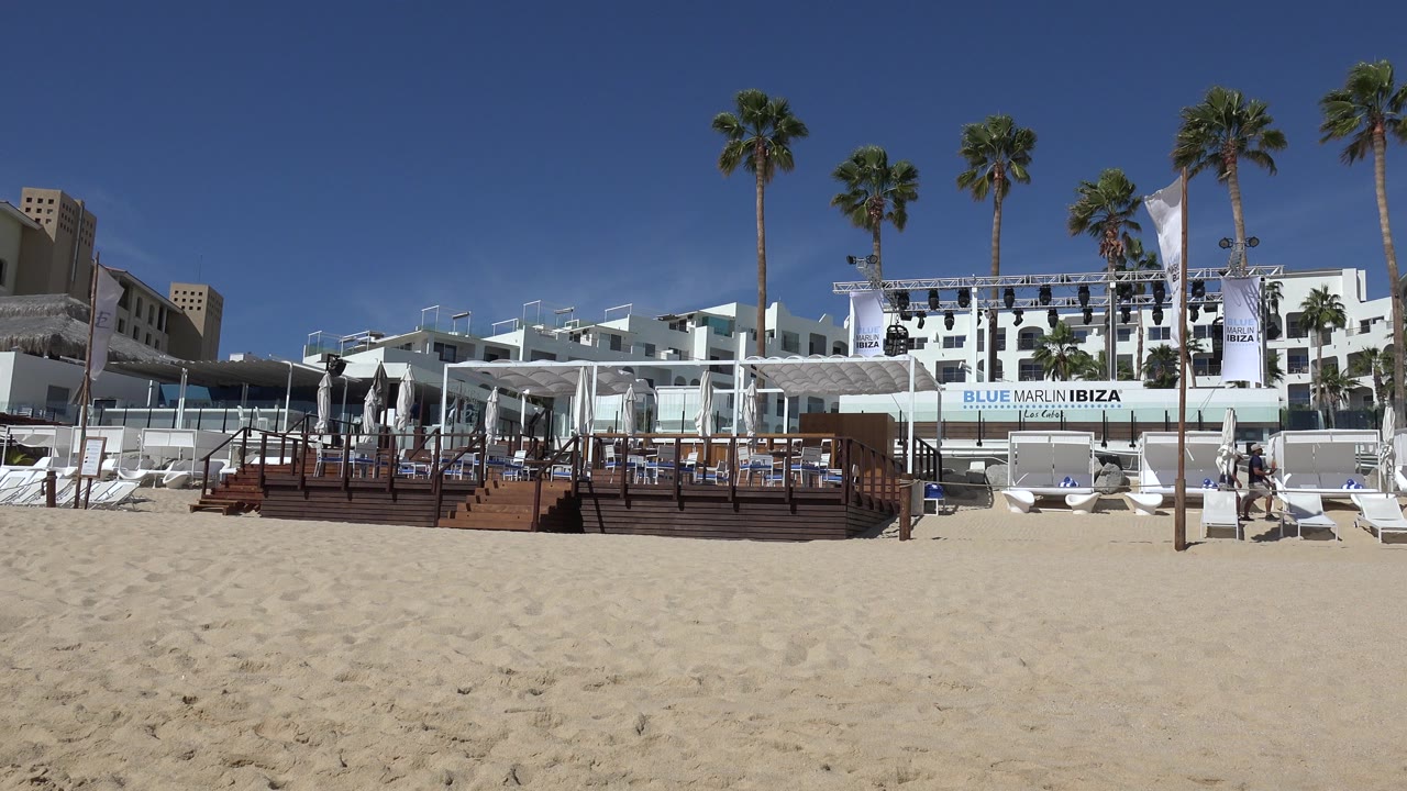 Cabo San Lucas beachfront resort exterior at the start of the clip, with pale sand, white buildings, palm trees, deck seating, lounge chairs, visible signage, and small distant people.